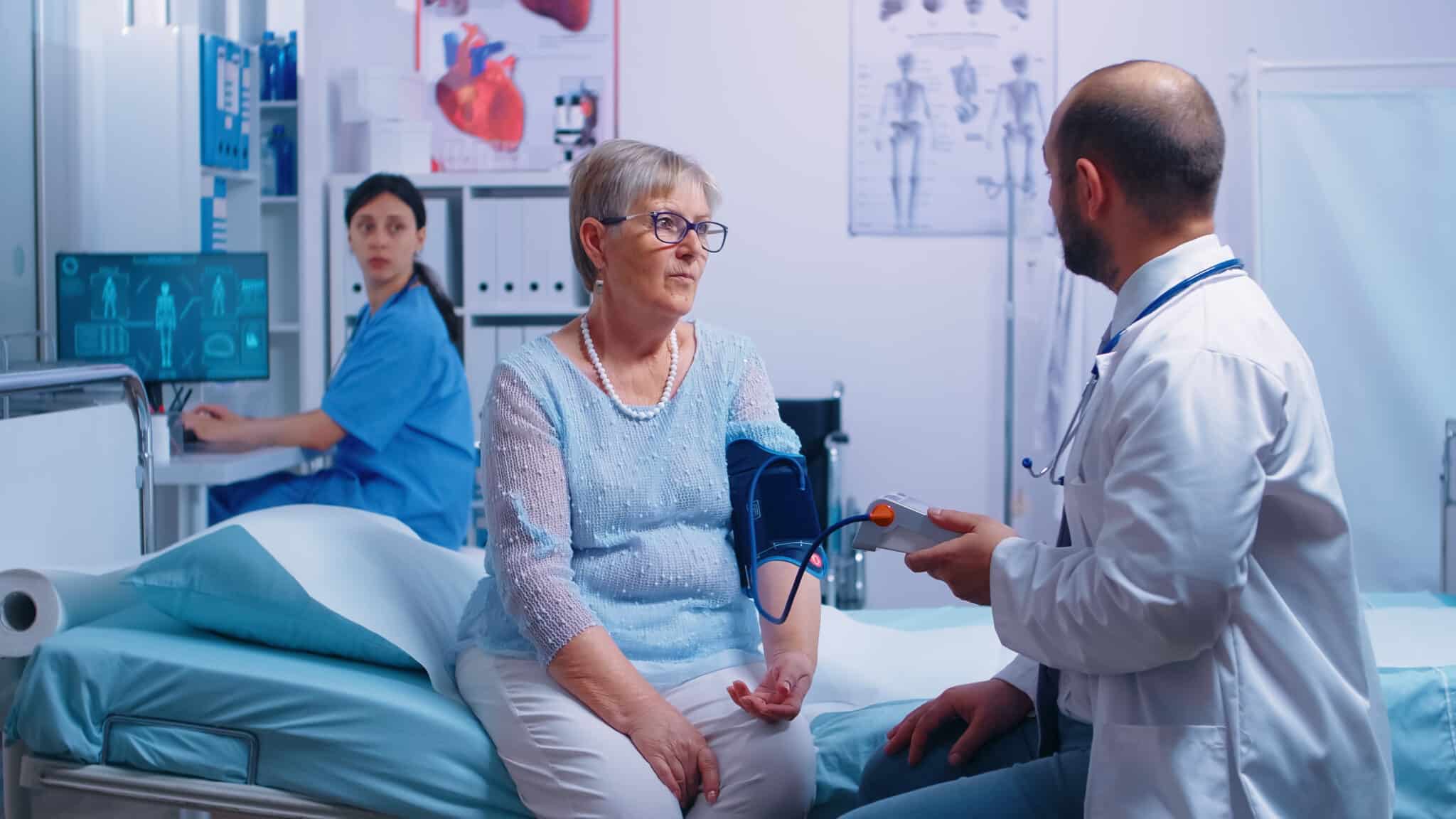 Physician treats patient in exam room, medical scribe is in the background documenting in the EHR.