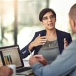 Accountant in a meeting uses laptop to show graphs; transcription plays a role in her data analysis.