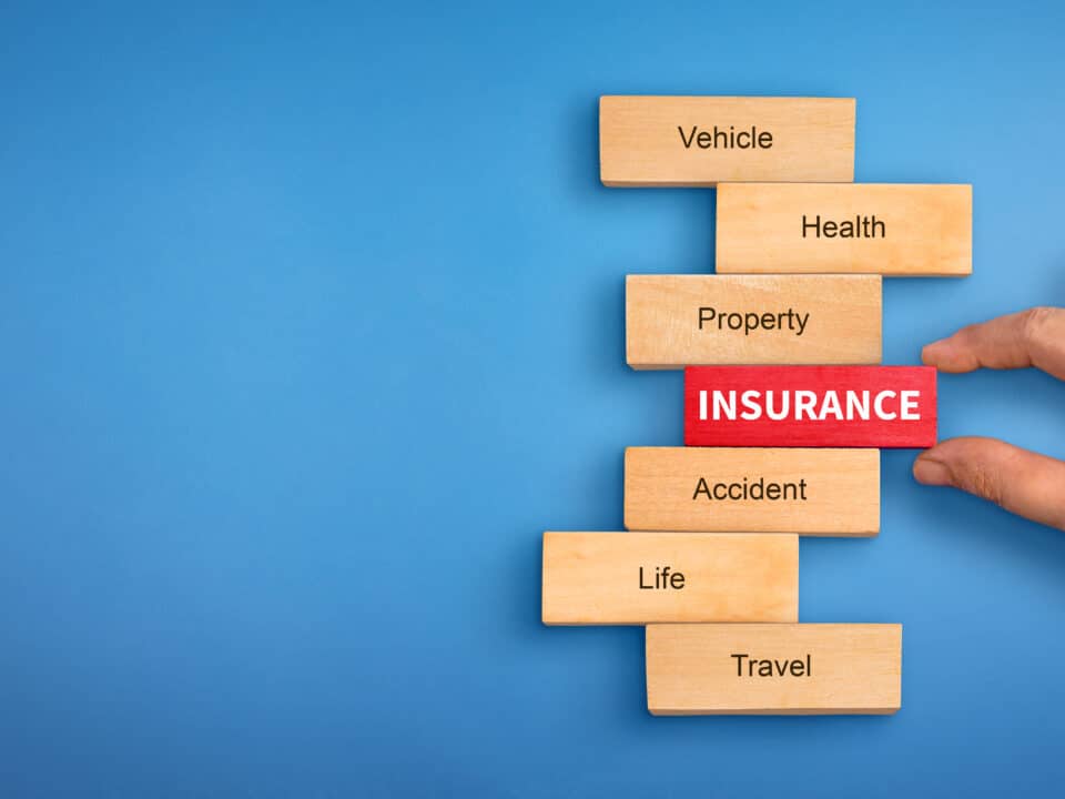 Hand holding a wooden 'Insurance' block, surrounded by blocks labeled with various insurance types.