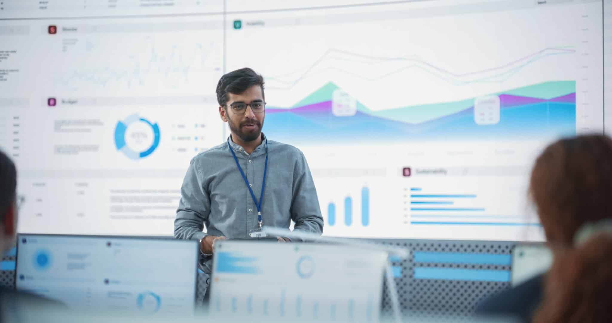 Indian computer scientist presents to diverse finance analysts with his tablet in a monitoring room.